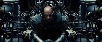 Movie still from “Riddick” (2013), directed by David Twohy – A man sitting on top of a chair wearing armor; Medium shot, Low angle