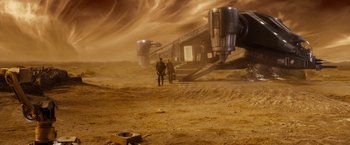 Movie still from “Riddick” (2013), directed by David Twohy – Two people walking on a dirt field near a building on top of a hill; Extreme Wide shot, High angle