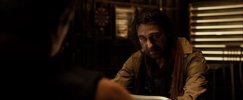 Movie still from “Riddick” (2013), directed by David Twohy – A man sitting at a table in front of a clock; Close Up shot, Over the shoulder angle