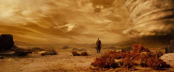 Movie still from “Riddick” (2013), directed by David Twohy – A man walking across a barren field with a cloudy sky; Extreme Wide shot, Over the shoulder angle