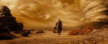 Movie still from “Riddick” (2013), directed by David Twohy – A man walking across a barren field holding a bag; Wide shot, Low angle