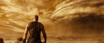 Movie still from “Riddick” (2013), directed by David Twohy – A man standing in front of a large storm cloud; Wide shot, Low angle