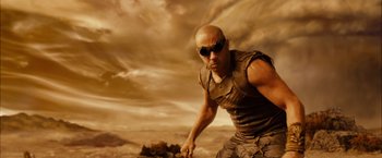 Movie still from “Riddick” (2013), directed by David Twohy – A bald man with sunglasses and a vest is standing in front of a cloudy sky; Medium shot, Low angle