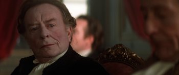 Movie still from “Ridicule” (1996), directed by Patrice Leconte – An older woman sitting in a chair with a man sitting in the background; Close Up shot, Over the shoulder angle