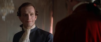 Movie still from “Ridicule” (1996), directed by Patrice Leconte – A man wearing a suit and tie smiling for the camera; Close Up shot, Over the shoulder angle