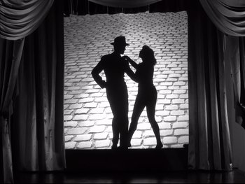 Movie still from “Rififi” (1955), directed by Jules Dassin – A man and a woman dancing on a stage in front of a brick wall; Wide shot, Low angle