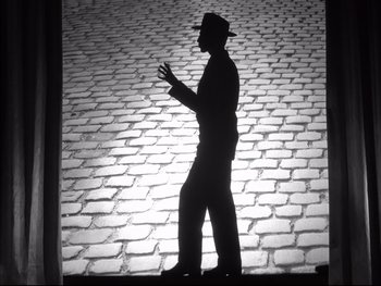 Movie still from “Rififi” (1955), directed by Jules Dassin – A man in a fedora standing on a cobblestone street; Wide shot, Low angle