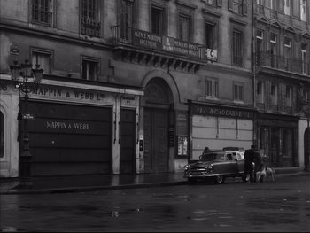 Movie still from “Rififi” (1955), directed by Jules Dassin – An old car parked in front of a building on a rainy day; Extreme Wide shot, High angle