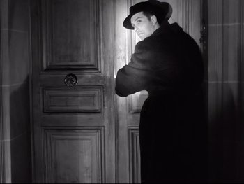 Movie still from “Rififi” (1955), directed by Jules Dassin – A man standing in front of a door wearing a hat; Medium shot, Low angle