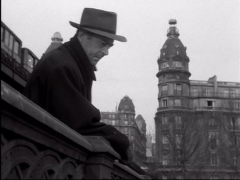 Movie still from “Rififi” (1955), directed by Jules Dassin – An older man sitting on a railing in front of some buildings; Medium shot, Low angle
