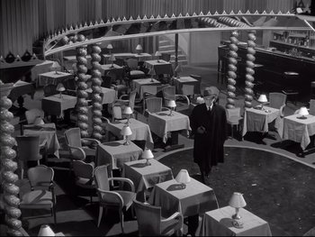 Movie still from “Rififi” (1955), directed by Jules Dassin – A man standing in front of a room filled with tables and chairs; Wide shot, High angle