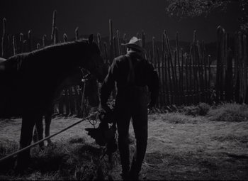 Movie still from “Rio Grande” (1950), directed by John Ford – A man is standing next to a horse in the dark; Wide shot, Low angle