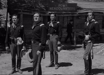 Movie still from “Rio Grande” (1950), directed by John Ford – A black and white photo of a group of men standing next to each other in front of a building; Wide shot, Low angle