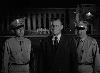 Movie still from “Riot in Cell Block 11” (1954), directed by Don Siegel – A black and white photo of a man and two men in uniform; Medium shot, Low angle