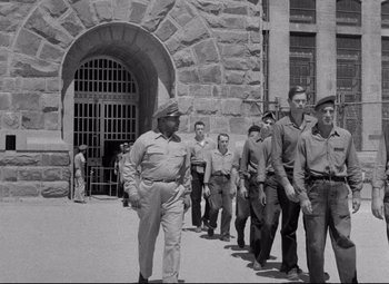 Movie still from “Riot in Cell Block 11” (1954), directed by Don Siegel – A black and white photo of men walking in front of a building; Wide shot, High angle