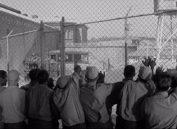 Movie still from “Riot in Cell Block 11” (1954), directed by Don Siegel – A black and white photo of a group of people looking over a chain link fence; Wide shot, High angle
