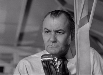 Movie still from “Riot in Cell Block 11” (1954), directed by Don Siegel – An older man holding a microphone in front of a wall; Close Up shot, Low angle