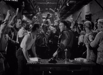 Movie still from “Riot in Cell Block 11” (1954), directed by Don Siegel – A group of men standing around a table; Medium shot, High angle