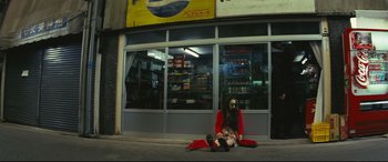 Movie still from “Ritual” (2000), directed by Hideaki Anno – A person sitting on the ground outside a store; Wide shot, Low angle