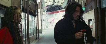 Movie still from “Ritual” (2000), directed by Hideaki Anno – A person standing on a sidewalk near a building; Medium shot, Low angle