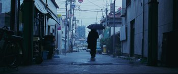 Movie still from “Ritual” (2000), directed by Hideaki Anno – A person with an umbrella walking down a street; Wide shot, Low angle