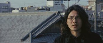 Movie still from “Ritual” (2000), directed by Hideaki Anno – A person standing on top of a building with buildings in the background; Close Up shot, Low angle