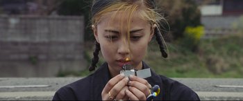 Movie still from “Ritual” (2000), directed by Hideaki Anno – A young girl is lighting a cigarette in her mouth; Close Up shot, High angle