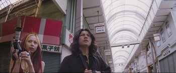 Movie still from “Ritual” (2000), directed by Hideaki Anno – A man with long hair and glasses standing in an alley way; Medium shot, Low angle