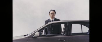 Movie still from “Ritual” (2000), directed by Hideaki Anno – A man standing in the driver's seat of a car; Medium shot, Low angle