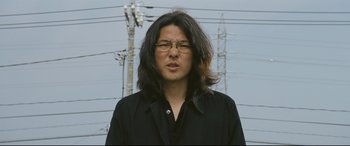 Movie still from “Ritual” (2000), directed by Hideaki Anno – A man with long black hair wearing glasses and a black shirt; Close Up shot, Low angle
