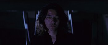 Movie still from “Ritual” (2000), directed by Hideaki Anno – A man with long black hair wearing glasses and a black shirt; Close Up shot, Low angle