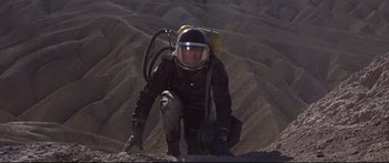Movie still from “Robinson Crusoe on Mars” (1964), directed by Byron Haskin – A man in a black suit and helmet is climbing up a hill; Wide shot, Overhead angle