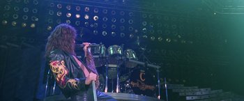 Movie still from “Rock Star” (2001), directed by Stephen Herek – A man sitting on top of a chair holding a microphone; Wide shot, High angle