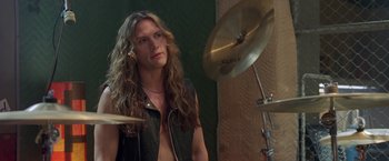 Movie still from “Rock Star” (2001), directed by Stephen Herek – A man with long curly hair is playing a drum set; Close Up shot, Over the shoulder angle