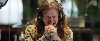 Movie still from “Rock Star” (2001), directed by Stephen Herek – A man with long hair wearing headphones is singing into a microphone; Close Up shot, Overhead angle