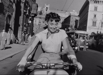 Movie still from “Roman Holiday” (1953), directed by William Wyler – A woman riding a motorcycle down a street; Medium shot, Low angle