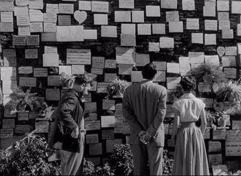Movie still from “Roman Holiday” (1953), directed by William Wyler – A group of people standing next to a wall covered in notes; Wide shot, Over the shoulder angle