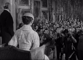 Movie still from “Roman Holiday” (1953), directed by William Wyler – An old photo of a woman sitting in front of an audience; Wide shot, Over the shoulder angle