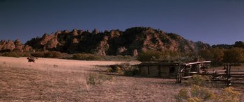 Movie still from “Romancing the Stone” (1984), directed by Robert Zemeckis – An old cabin sits in the middle of a field near a mountain range; Extreme Wide shot, High angle