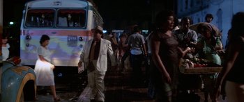 Movie still from “Romancing the Stone” (1984), directed by Robert Zemeckis – A group of people walking down a sidewalk at night near a bus; Wide shot, Over the shoulder angle