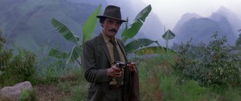 Movie still from “Romancing the Stone” (1984), directed by Robert Zemeckis – A man holding a gun while standing in a field; Medium shot, High angle