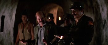 Movie still from “Romancing the Stone” (1984), directed by Robert Zemeckis – A group of men standing around each other holding candles; Medium shot, Over the shoulder angle