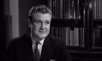 Movie still from “Room at the Top” (1958), directed by Jack Clayton – Black and white photograph of a man in a suit and tie in front of a bookshelf; Close Up shot, Low angle
