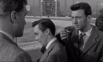 Movie still from “Room at the Top” (1958), directed by Jack Clayton – A man is smoking a cigarette while two other men look on; Medium shot, Over the shoulder angle