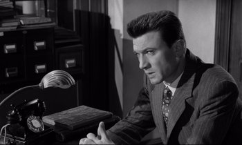 Movie still from “Room at the Top” (1958), directed by Jack Clayton – A black and white photo of a man sitting at a desk; Medium shot, Low angle