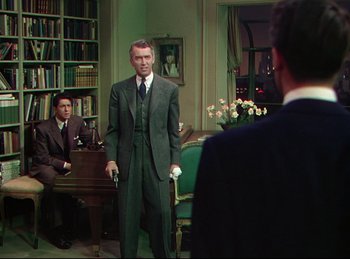 Movie still from “Rope” (1948), directed by Alfred Hitchcock – A man in a suit and tie standing in front of two other men in a room; Medium shot, Over the shoulder angle