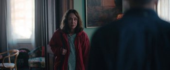 Movie still from “Rose” (2022), directed by Niels Arden Oplev – A woman wearing a red jacket standing in front of a painting; Medium shot, Over the shoulder angle