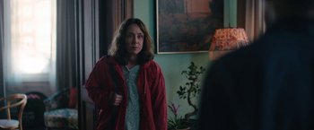 Movie still from “Rose” (2022), directed by Niels Arden Oplev – A woman wearing a red jacket standing in front of a painting; Medium shot, Over the shoulder angle