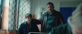 Movie still from “Rose” (2022), directed by Niels Arden Oplev – A man standing next to a young boy in a room; Medium shot, Over the shoulder angle