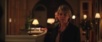 Movie still from “Rose” (2022), directed by Niels Arden Oplev – A woman standing next to a table in a room; Medium shot, Over the shoulder angle